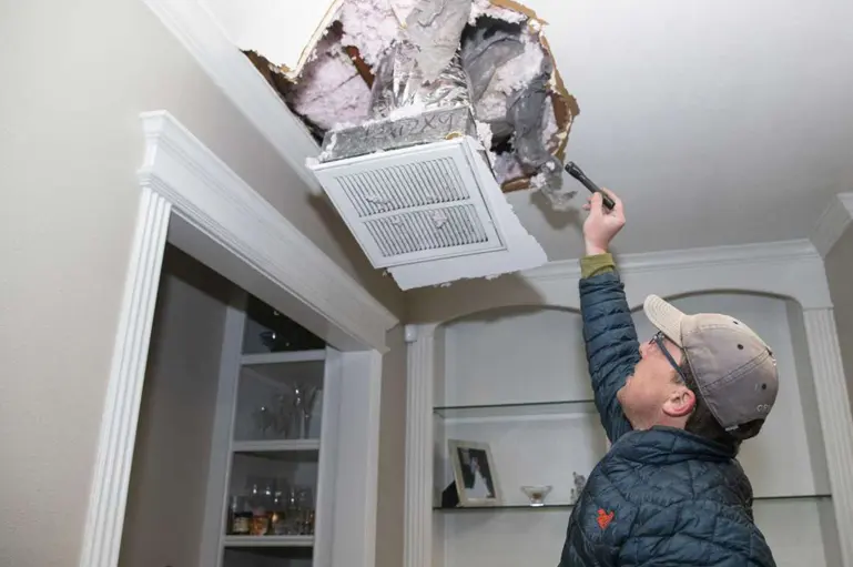 Man in a cap looking into an opened ceiling beside an air vent